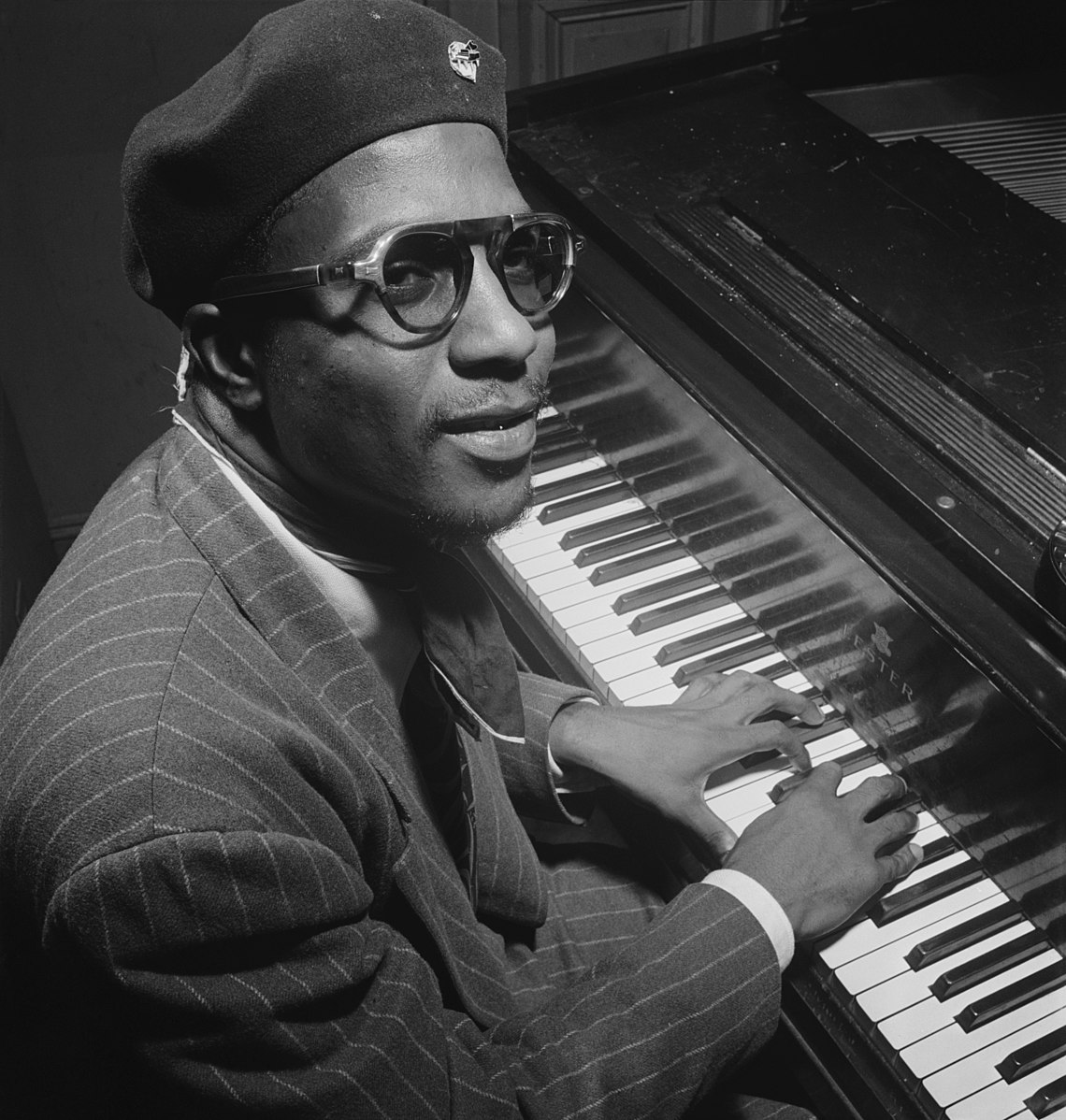 Man playing piano and looking up at camera in hat and glasses.
