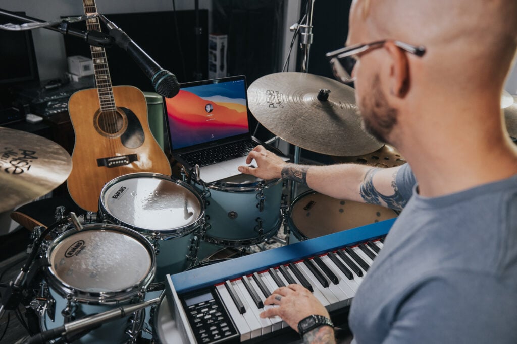 Over the shoulder shot of man with glasses using laptop while playing keyboard with drums and guitar.