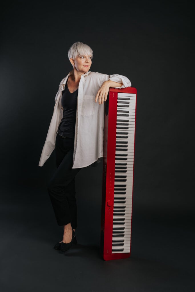 Woman with short platinum hair in white airy blouse leaning casually on a vertical red keyboard.