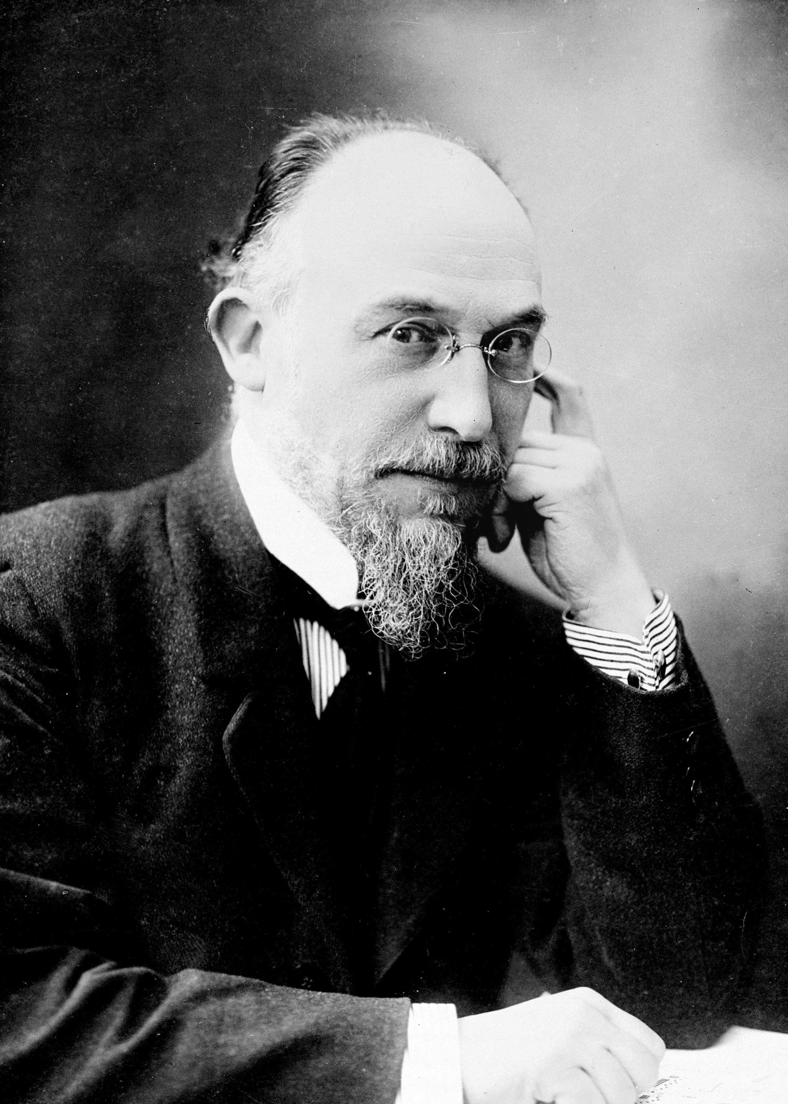 Black and white photograph of Erik Satie, composer of Gymnop&eacute;die No. 1 on piano. Middle-aged man in suit with thoughtful expression, scraggly beard, and glasses.