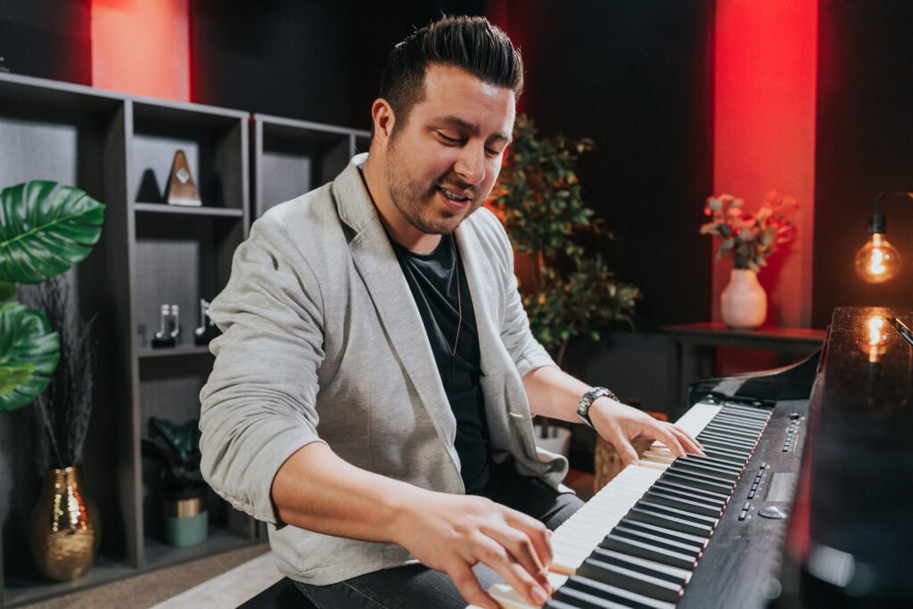 Man with dark hair in light grey suit jacket playing grand piano in studio.