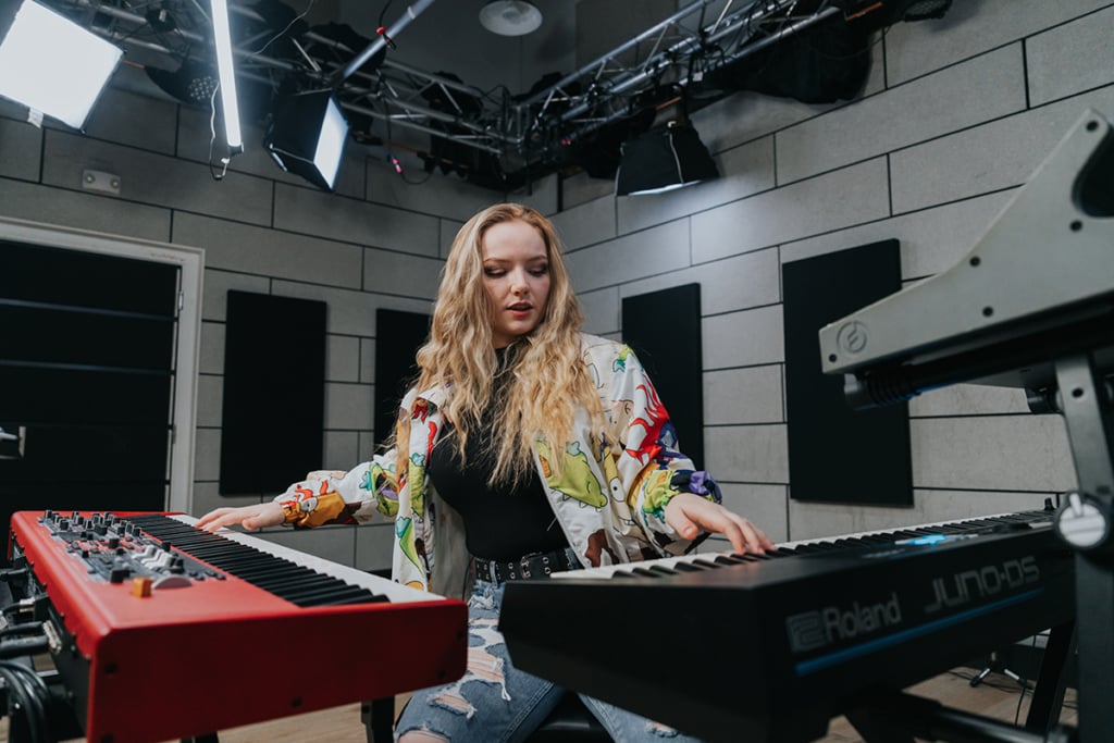 Woman with long blonde hair and cartoon character hoodie playing multiple keyboards at the same time.