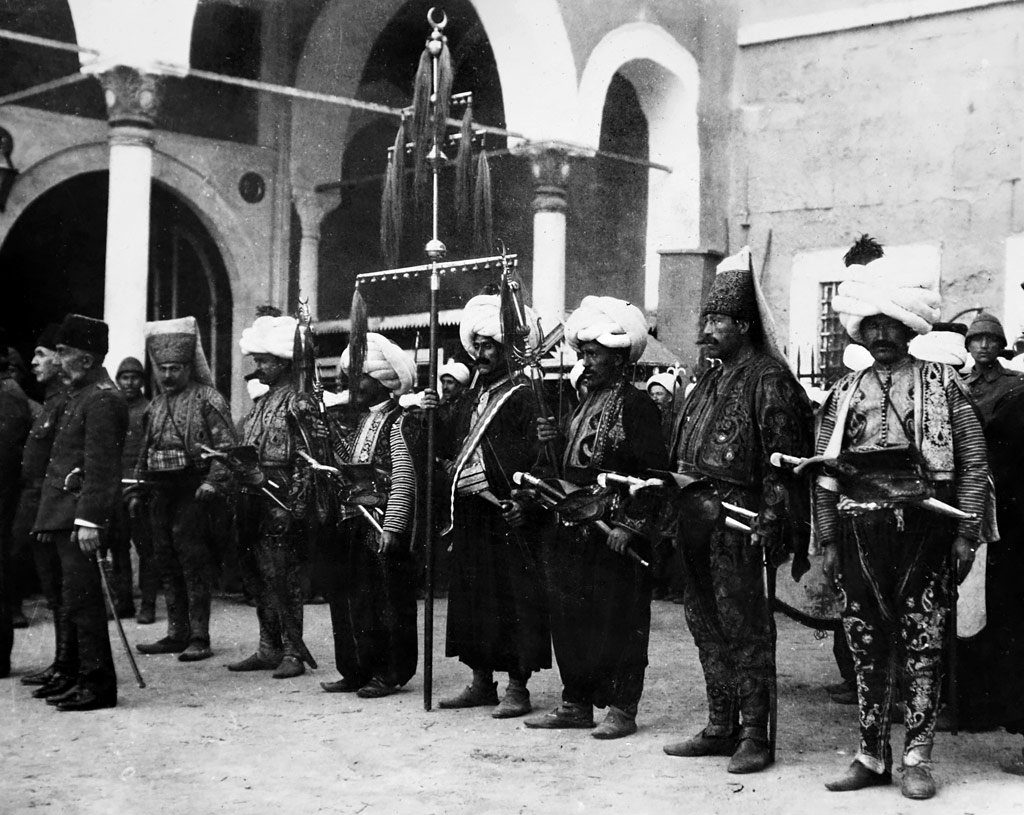 Black and white photo of men in traditional Turkish clothing and turbans holding instruments while standing.